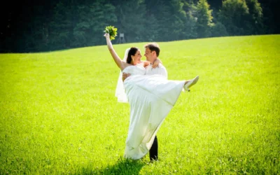 Brautpaar in Wiese, Braut wird hochgehoben, lachend. Hochzeitsfoto nahe Wolfratshausen. Romantischer Moment im Freien.