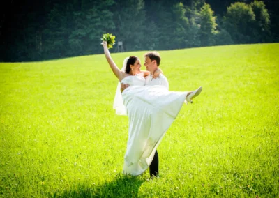 Brautpaar in Wiese, Braut wird hochgehoben, lachend. Hochzeitsfoto nahe Wolfratshausen. Romantischer Moment im Freien.
