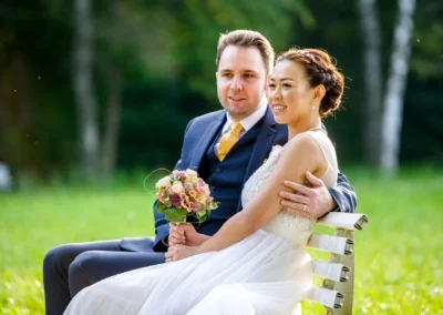 Brautpaar sitzt auf einer Bank in Fischbachau, Braut mit Blumenstrauss, Hochzeitsfotografie im Freien.