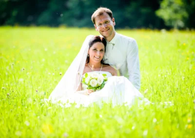 Brautpaar sitzt lächelnd im Gras auf Sprengenöder Alm nahe Wolfratshausen. Hochzeitsfotografie mit Blumenstrauss bei sommerlicher Hochzeit.