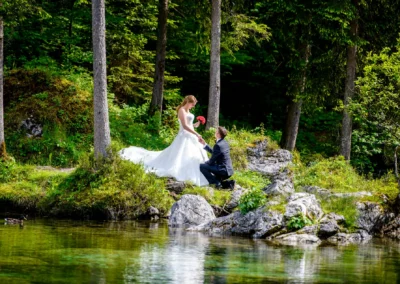 Brautpaarfoto im Grünen, nahe Badersee. Braut im weißen Kleid mit Blumenstrauß, Bräutigam kniet am Ufer. Hochzeitsreportage.
