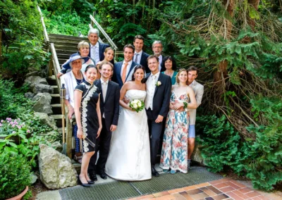 Gruppenfoto bei Hochzeit nahe Wolfratshausen, Brautpaar und Gaeste in eleganter Kleidung, gruen bewachsene Kulisse.