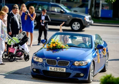 Brautpaar im blauen Cabrio bei Hochzeit am Starnberger See, von Familie und Freunden fotografiert. Blumenschmuck.