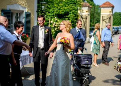 Brautpaar lacht mit Gaesten bei Hochzeit in Moosach. Hochzeitsfoto einer sommerlichen Feier unter freiem Himmel.