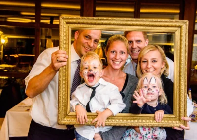 Fröhliche Hochzeitsgesellschaft beim Fotoshooting mit Rahmen im Paulaner Seegarten, Kinder mit Gesichtsbemalungen.