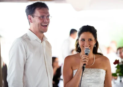 Brautpaar lacht bei Hochzeit mit Mikrofon, Hochzeitsfoto auf der Sprengenöder Alm nahe Wolfratshausen, festliche Stimmung, Hochzeitsfotograf.