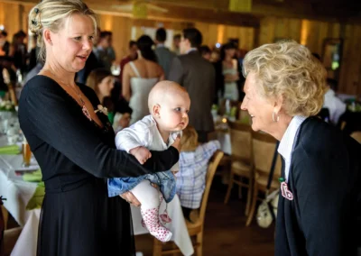Ein Baby wird bei einer Hochzeitsfeier in Sauerlach von einer Frau gehalten und mit einer aelteren Dame interagiert.