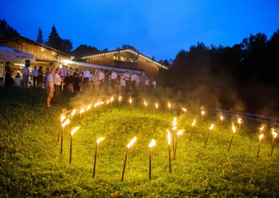 Gäste stehen bei einer Hochzeit auf der Sprengenöder Alm bei Wolfratshausen, umgeben von Fackeln im Gras. Hochzeitsfoto in Oberbayern.