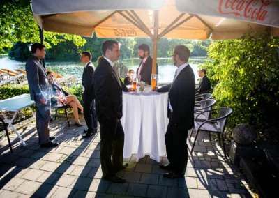 Gaste in Anzug bei Hochzeit am Kleinhesseloher See. Entspannte Unterhaltung im Freien bei einem Stehtisch.