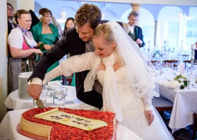 Brautpaar schneidet Hochzeitstorte in Tegernsee, umgeben von Gästen. Hochzeitsreportage zeigt festliche Stimmung.