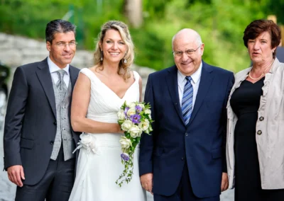 Brautpaar mit Familie laechelt bei Hochzeit am Leeberghof, Brautstrauss aus weissen und lila Blumen.