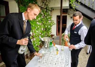 Hochzeitsfoto vom Sektempfang am Eibsee Hotel. Zwei Maenner im Anzug schenken Sekt in Glaeser ein.