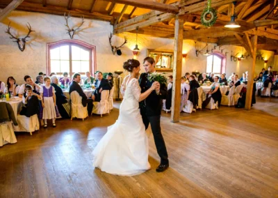 Brautpaar tanzt in rustikaler Scheune auf Hochzeit im Kreis Rosenheim, Gaeste sitzen an runden Tischen.