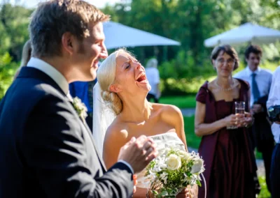 Brautpaar lacht bei einer Hochzeitsfeier in Oberambach, umgeben von Gaesten im Freien. Hochzeitsfotografie.