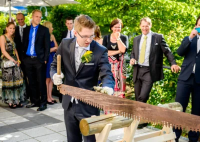 Bräutigam sägt Holz bei Hochzeitsfeier im Vier Jahreszeiten Starnberg, umringt von Gästen. Hochzeitsfotografie.