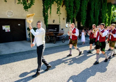 Mann mit Besen und Musikanten bei Hochzeitszug nahe Rosenheim. Hochzeitsreportage im Landgasthof Stahuber Thal.