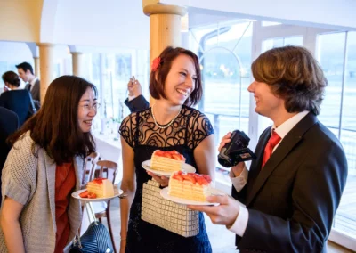 Gäste lachen mit Kuchen bei einer Hochzeit am Tegernsee, Hochzeitsfoto zeigt fröhliche Szene in einem Café.