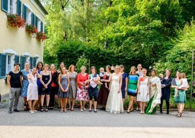 Gruppe von Hochzeitsgaesten in Moosach vor einem gelben Haus mit Fensterlaeden und Bluemendekoration.