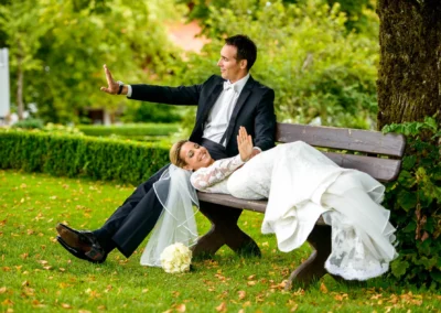 Brautpaar lacht auf Parkbank im Grünen bei Hochzeits-Portrait nahe Tegernsee. Elegantes Hochzeitsfoto voller Freude.
