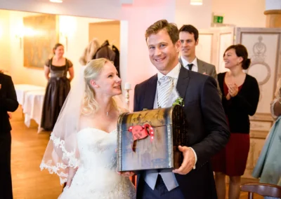 Brautpaar lacht mit Schatztruhe bei der Hochzeitsfeier in Tegernsee, festgehalten von einem Hochzeitsfotografen.