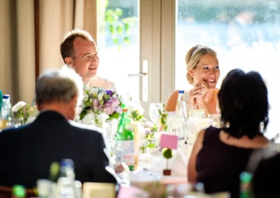 Brautpaar lacht waehrend Hochzeit in Schwabing. Blumenschmuck und Gaesten im Hintergrund einer Hochzeitsfotoreportage.