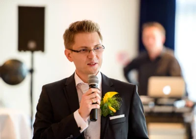 Mann im Anzug haelt Rede bei Hochzeit am Starnberger See, Hochzeitsfoto mit Mikrofon und gelber Blume im Anstecker.