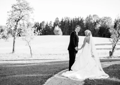 Brautpaar steht auf Wiese bei Schlossgut Oberambach, laechelnd, elegant gekleidet, umgeben von Baeumen.