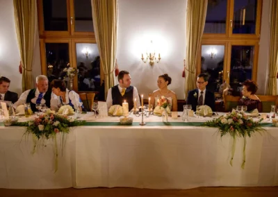 Hochzeitsfoto in Fischbachau, Brautpaar mit Gaesten an festlich gedecktem Tisch mit Blumenschmuck und Kerzen.