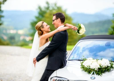 Brautpaarshooting in Waakirchen mit Brautstrauss, Braut umarmt Bräutigam vor Auto, Hochzeitsfotografie am Tegernsee.