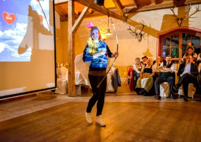 Frau mit Pfeil und Bogen bei Hochzeit im Hofgut zur Muehle. Fröhliche Gäste im Hintergrund. Hochzeitsreportage.