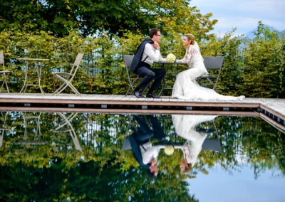 Brautpaar am Tisch, spiegelnd im Wasser, umgeben von Gruen bei Margarethenhof. Hochzeits-Foto nahe Tegernsee.