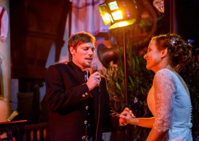 Bräutigam singt zum Mikrofon gerichtet, Braut lächelt, Hochzeit im Hofgut zur Mühle, festliche Beleuchtung.