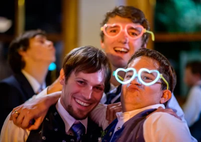 Drei lachende Gaeste auf einer Hochzeitsparty mit leuchtenden Herzbrillen in Hofgut zur Muehle. Hochzeitsfotografien.