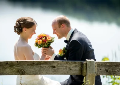 Brautpaar bei Hochzeit in irschenberg, sitzt auf Holzbank, lacht und haelt Blumenstrauss. Hochzeitsreportage.