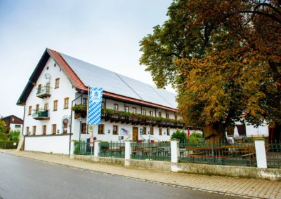 Großes weißes Gebäude mit roten Ziegeldach und bayerischer Fahne. Baum und Straße im Vordergrund. Augustlhof in Pfettrach bei Landshut.