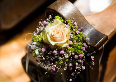 Blumenschmuck mit roser Rose und lila Blumen auf Holzbank bei kirchlicher Trauung in Schloss Aufhausen nahe Muenchen.