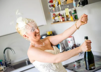 Braut im Brautkleid lacht beim Oeffnen einer Champagnerflasche in Kueche, Hochzeitsfoto in Bad Toelz.