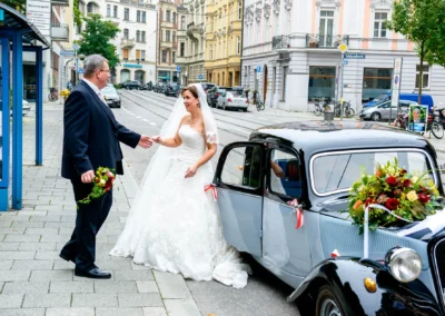 Braut steigt bei ihrer Hochzeit in Muenchen nahe Isar aus einem Oldtimer. Hochzeitsfotograf dokumentiert kirchliche Trauung.