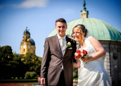 Brautpaar im Hofgarten Muenchen, laechelnd vor Pavillon, Hochzeitsfotografie mit rotem Brautstrauss, sonniger Tag.