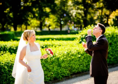 Brautpaarfoto im Hofgarten Muenchen, Braut mit Blumenstrauss laechelt den Bräutigam an, der Seifenblasen macht.