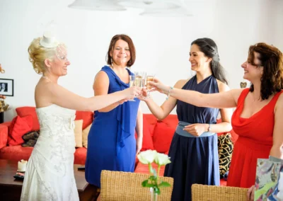 Braut in weissem Kleid stoesst mit drei Frauen im Wohnzimmer an, Hochzeitsfotografie in Bad Toelz, Oberbayern.