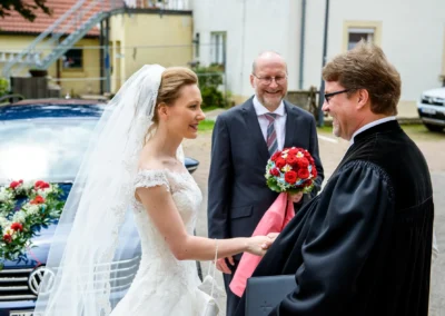 Braut in weissem Kleid schuettelt dem Geistlichen die Hand; Auto mit Blumenschmuck im Hintergrund in Ansbach.