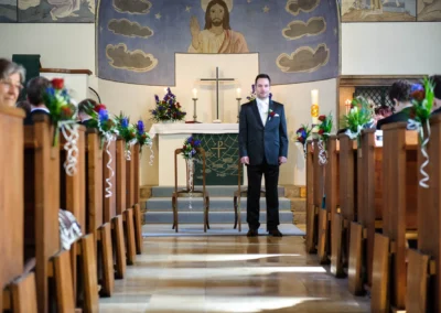 Bräutigam vor Altar in Christuskirche Gauting bei München. Hochzeitsfotografie zeigt kirchliche Zeremonie mit Blumen.