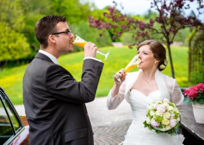 Brautpaar beim Anstossen mit Sekt in Aschbacher Hof, Hochzeitsfoto im Freien mit Blumenstrauss.