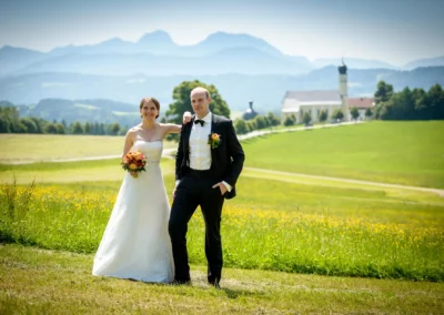 Brautpaar posiert auf Wiese in Irschenberg, Kirche St. Johann-Baptist im Hintergrund, Hochzeit Hochzeitsfotografie.