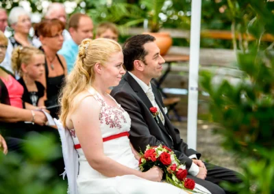 Brautpaar bei freier Trauung in Röhrmoos mit roten Rosenstrauss, umgeben von Hochzeitsgaesten im Gruenen.