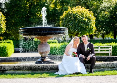 Brautpaar beim Hochzeitsshooting vor Brunnen im Hofgarten Muenchen, umgeben von gruenem Laub und Sonne.