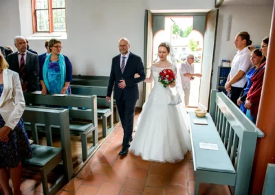 Braut mit Blumenstrauss schreitet mit Begleiter durch Kirche bei Hochzeit in Ansbach, Gaeste stehen auf Kirchbaenken.