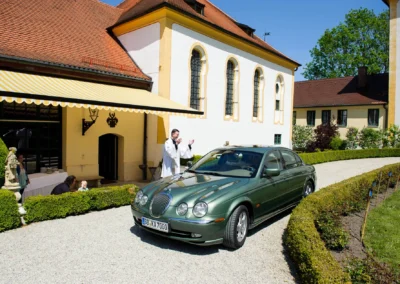 Priester segnet Auto vor der Kirche bei einer Hochzeit im Schloss Aufhausen nahe Muenchen. Hochzeitsreportage.