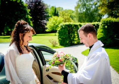 Braut trifft Priester mit Blumenstrauss vor kirchlicher Trauung im Schloss Aufhausen nahe Muenchen. Hochzeitsreportage.